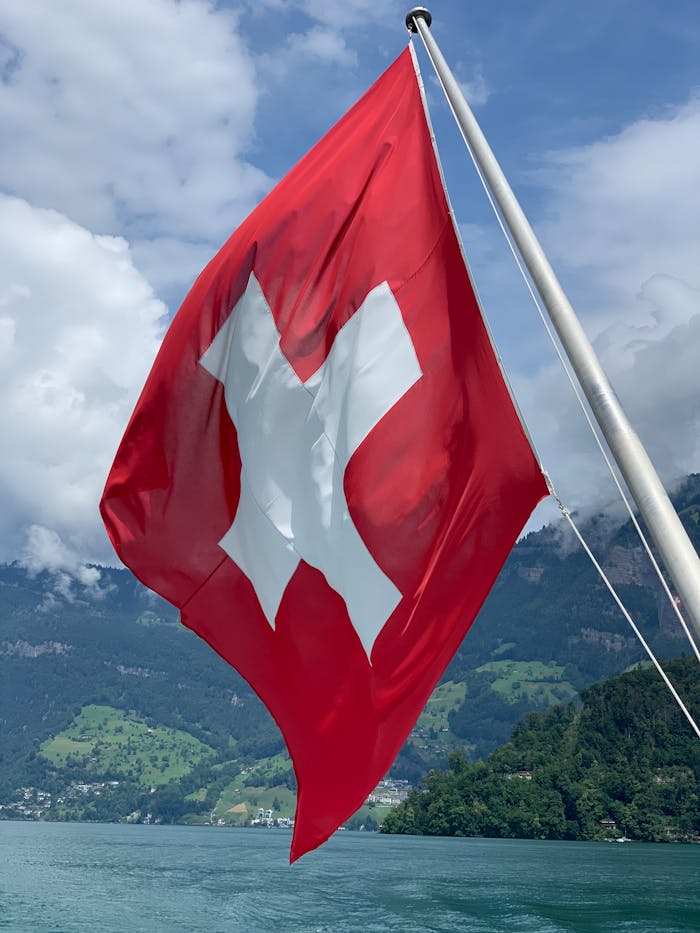 The Swiss flag waves against a scenic backdrop of a lake and mountains.