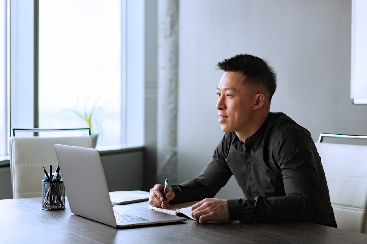 Asian male professional working at a desk in a modern office with a laptop and notepad.